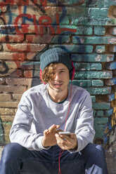 Portrait of young man with headphones and cell phone sitting in front of graffiti wall - AFVF00313