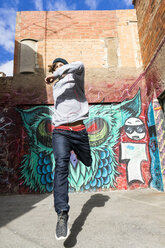 Junger Mann mit Kopfhörern, der vor einem Graffiti in die Luft springt - AFVF00330