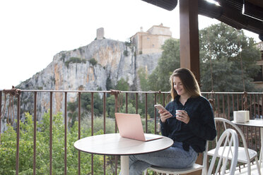 Spanien, Alquezar, lächelnde junge Frau mit Laptop auf Terrasse sitzend und mit Handy telefonierend - AFVF00335