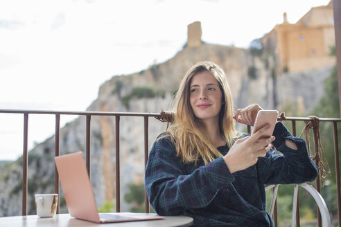 Spanien, Alquezar, Porträt einer entspannten jungen Frau mit Laptop und Mobiltelefon auf einer Terrasse sitzend - AFVF00337