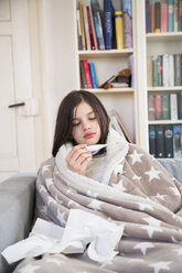 Sick girl sitting on the couch at home looking at clinical thermometer - LVF06799