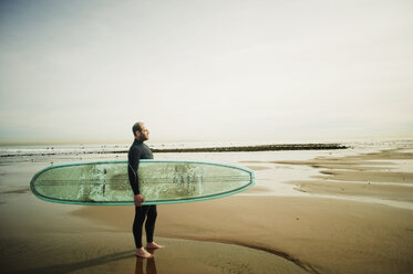 Seitenansicht eines Mannes mit Surfbrett, der wegschaut, während er auf Sand am Strand gegen den Himmel steht - CAVF11960