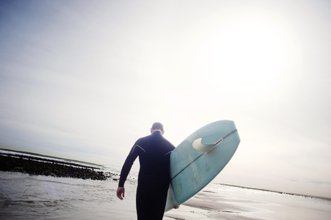 Rückansicht eines Mannes, der ein Surfbrett trägt, während er am Ufer gegen den Himmel läuft, lizenzfreies Stockfoto