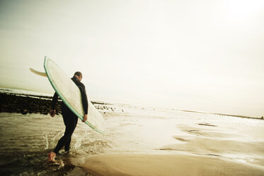 Rückansicht eines Mannes, der ein Surfbrett trägt, während er am Strand spazieren geht - CAVF11962