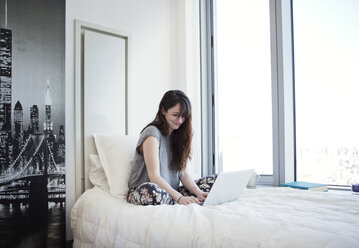 Lächelnde Frau, die einen Laptop benutzt, während sie zu Hause auf dem Bett sitzt - CAVF12010