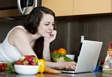 Frau benutzt einen Laptop, während sie sich zu Hause auf die Küchentheke stützt - CAVF12024