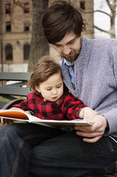 Man reading book while sitting with daughter on bench at park - CAVF12059