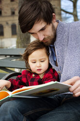Mann liest ein Buch und sitzt mit seiner Tochter auf einer Bank - CAVF12060
