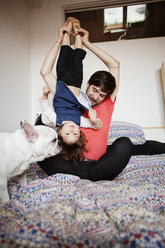 Father carrying daughter upside down while sitting by dog on bed - CAVF12063