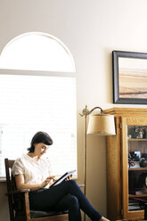 Ernste Frau, die ein digitales Tablet benutzt, während sie zu Hause am Bogenfenster sitzt - CAVF12275