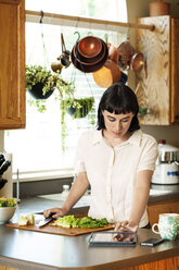 Frau liest digitales Tablet beim Schneiden von frischem Gemüse in der Küche zu Hause - CAVF12279