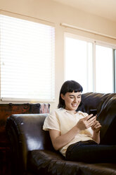 Smiling woman using phone while relaxing on sofa at home - CAVF12282