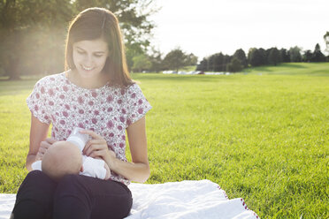 Lächelnde Mutter, die ihr Baby füttert, während sie auf einer Wiese im Park sitzt - CAVF12295