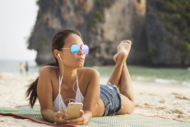 Frau hört Musik über ihr Smartphone, während sie auf einer Matte am Strand liegt - CAVF12378