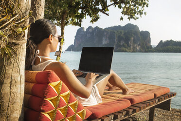 Woman using laptop computer while sitting on lounge chair against sea - CAVF12379