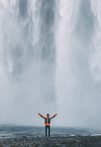 Mann steht mit ausgestreckten Armen vor dem Wasserfall Skogafoss in Island, lizenzfreies Stockfoto