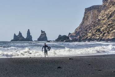 Mann mit Surfbrett zu Fuß am schwarzen Strand gegen den klaren Himmel an einem sonnigen Tag - CAVF12396