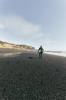 Rückansicht eines Mannes, der ein Surfbrett trägt, während er am schwarzen Strand gegen den Himmel an einem sonnigen Tag läuft - CAVF12402