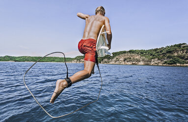 Shirtless man with surfboard jumping into sea against sky during sunny day - CAVF12414