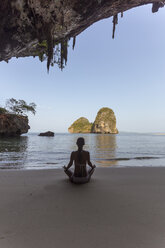 Rear view of woman in bikini meditating at beach seen through cave - CAVF12424