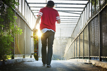 Rückansicht eines Mannes, der ein Skateboard trägt, während er über eine Brücke läuft - CAVF12455