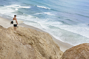 Mann trägt Surfbrett und steht auf einer Felsformation am Strand - CAVF12456