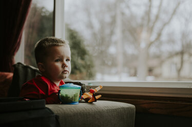 Thoughtful boy looking away while sitting on chair by window at home - CAVF12606