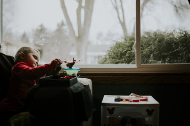 Boy playing with toy while sitting on chair by window at home - CAVF12607