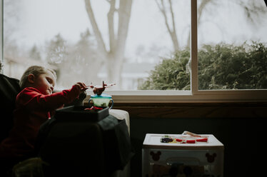 Junge spielt mit Spielzeug, während er zu Hause auf einem Stuhl am Fenster sitzt - CAVF12607