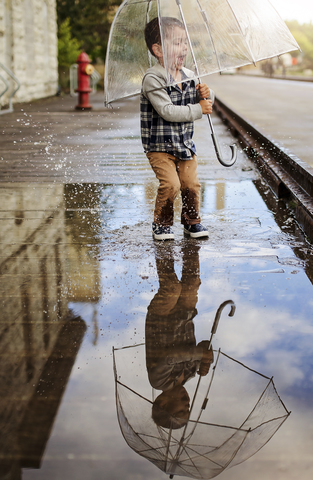 Verspielter Junge mit Regenschirm springt auf Pfütze an Straße, lizenzfreies Stockfoto