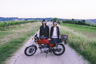 Portrait of young couple in crash helmets and flying goggles standing behind motorcycle at country road against clear sky - CAVF12654