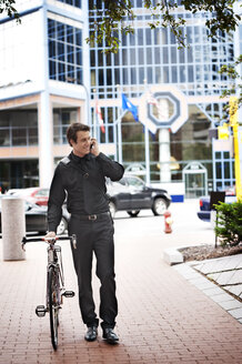 Businessman talking on phone while walking with bicycle on footpath in city - CAVF13122