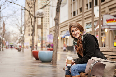 Glückliche Frau hält ein Einwegglas, während sie auf einer Bank in der Stadt sitzt - CAVF13181