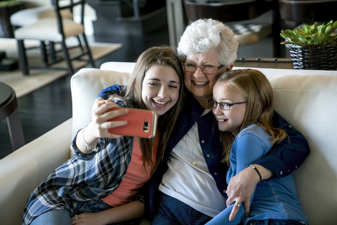Hohe Winkel Ansicht der glücklichen Großmutter und Enkelinnen nehmen Selfie durch Smartphone, während auf dem Sofa sitzen, lizenzfreies Stockfoto