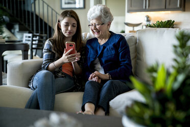 Enkelin zeigt der Großmutter ihr Smartphone, während sie zu Hause auf dem Sofa sitzt - CAVF13265