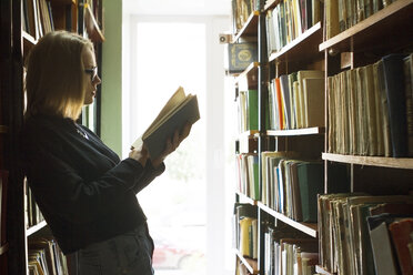 Side view of woman reading book while leaning on bookshelf - CAVF13295