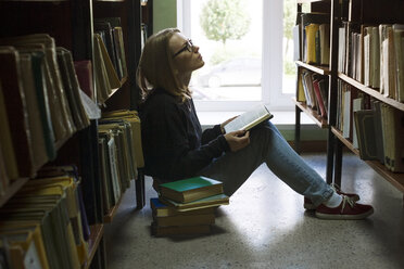 Frau hält ein Buch in der Hand und schaut auf, während sie in einer Bibliothek sitzt - CAVF13296