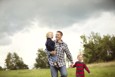 Smiling father with sons on grassy field against sky - CAVF13761