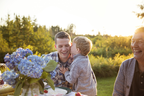 Junge küsst Vater am Tisch beim Picknick gegen den Himmel, lizenzfreies Stockfoto