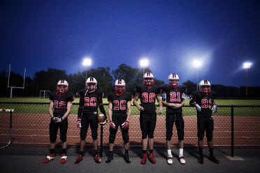 Male American football team standing by fence at illuminated field - CAVF13861