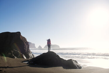 Rear view of woman standing on rock at shore against sky - CAVF13914