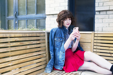 Frau benutzt Smartphone, während sie auf einer Bank an der Wand sitzt - CAVF14177