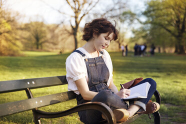 Frau mit gekreuzten Beinen beim Schreiben auf einer Parkbank sitzend - CAVF14184