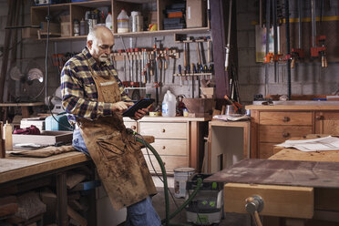 Ein älterer Handwerker benutzt ein digitales Tablet, während er in der Werkstatt steht - CAVF14390