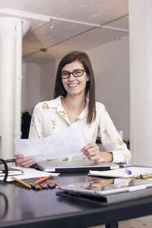 Selbstbewusst lächelnde Geschäftsfrau, die ein Dokument hält, während sie am Tisch sitzt - CAVF14403