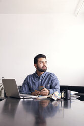 Thoughtful man with laptop and mobile phone sitting at table against wall - CAVF14405