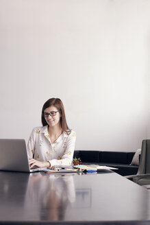 Selbstbewusste Geschäftsfrau, die einen Laptop benutzt, während sie an einem Tisch an der Wand sitzt - CAVF14408