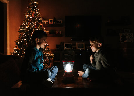 Brothers with illuminated lantern sitting by Christmas tree at home - CAVF14555