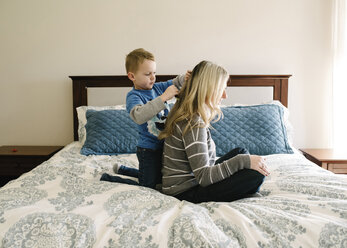 Niedlicher Sohn kämmt die Haare seiner Mutter, während er auf dem Bett vor der Wand kniet - CAVF14586