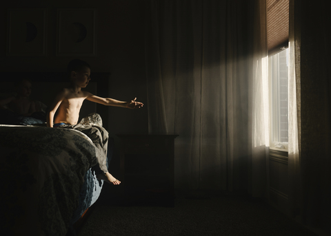 Hemdloser Junge versucht, das Sonnenlicht zu berühren, während er auf dem Bett in der Dunkelkammer sitzt, lizenzfreies Stockfoto